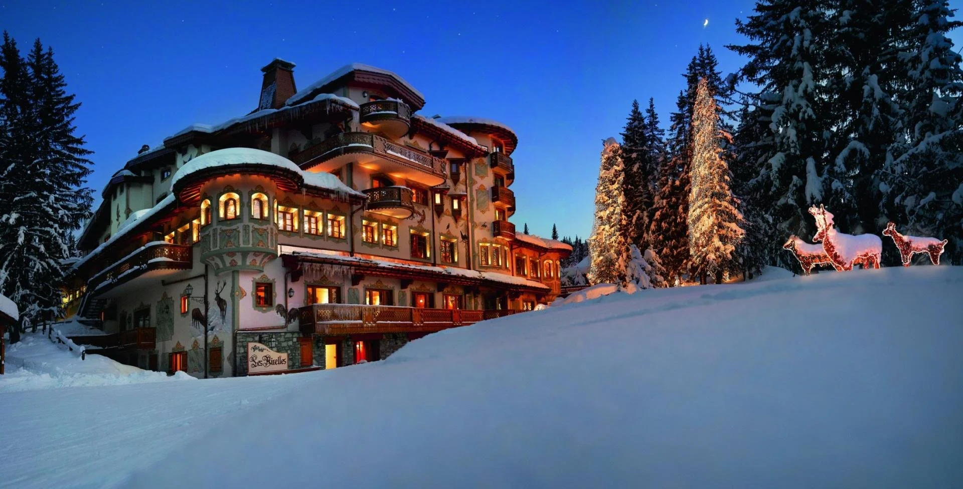 A large snow-covered chalet-style building with warm lights glowing from its windows, surrounded by snow-laden trees. There are illuminated reindeer figures on the snow in front of the trees, under a twilight sky with a crescent moon and stars.