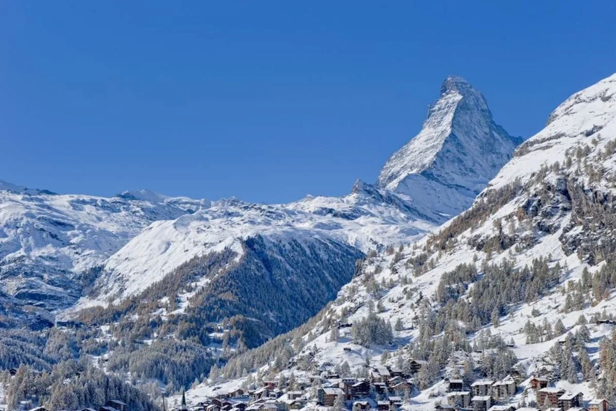 Snow-covered mountains with Zermatt village at their base and the Matterhorn mountain peak in the background under a clear blue sky.