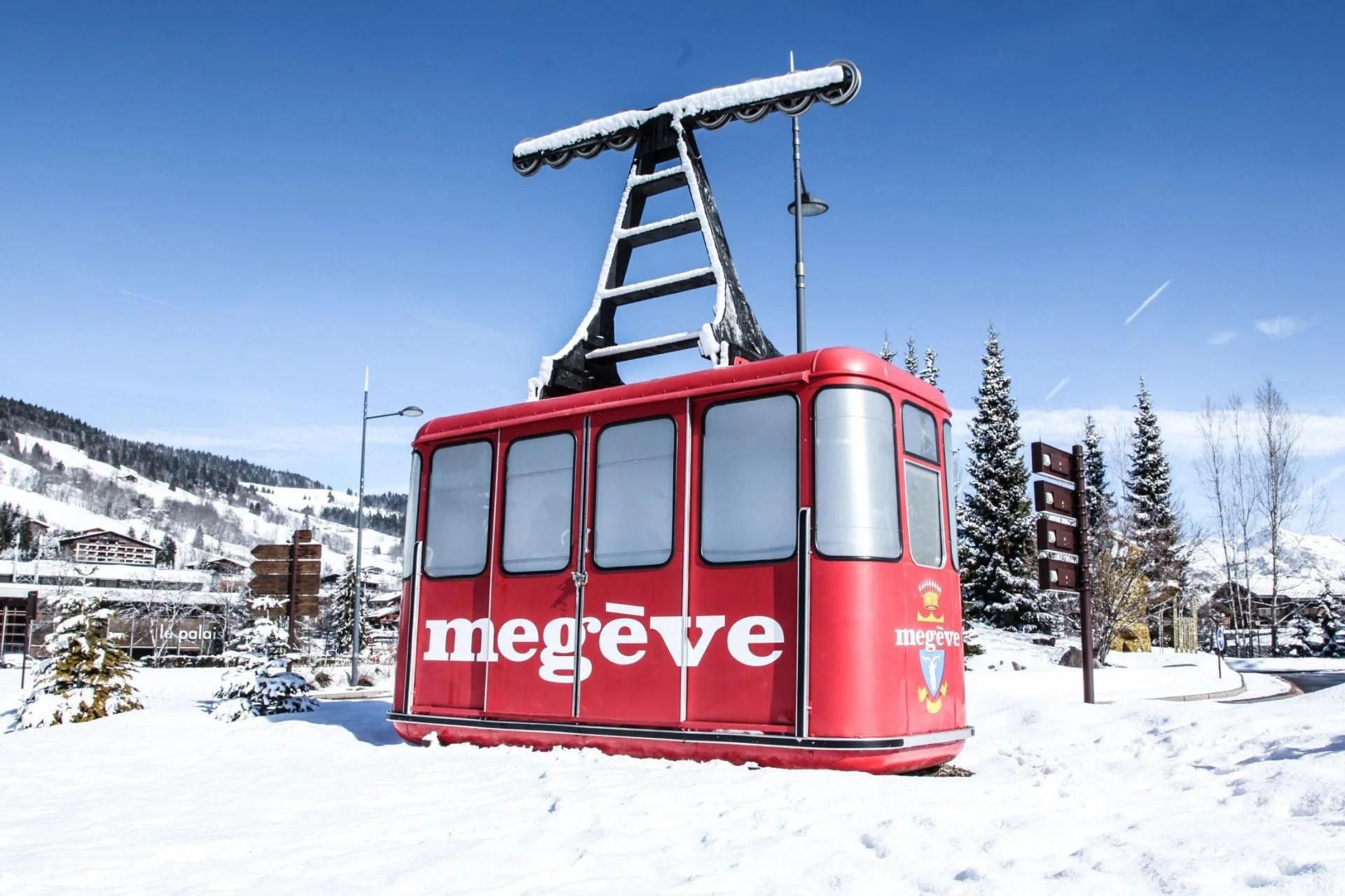 Red Megeve cable car with snow on its roof, set in a snowy landscape with pine trees and mountains in the background.