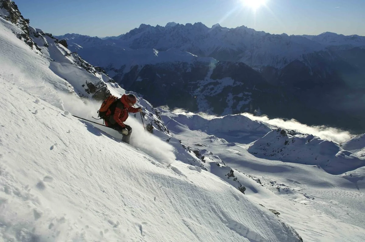 A skier in red gear descending a steep snowy mountain slope in Verbier with mountainous terrain in the background under a clear blue sky.