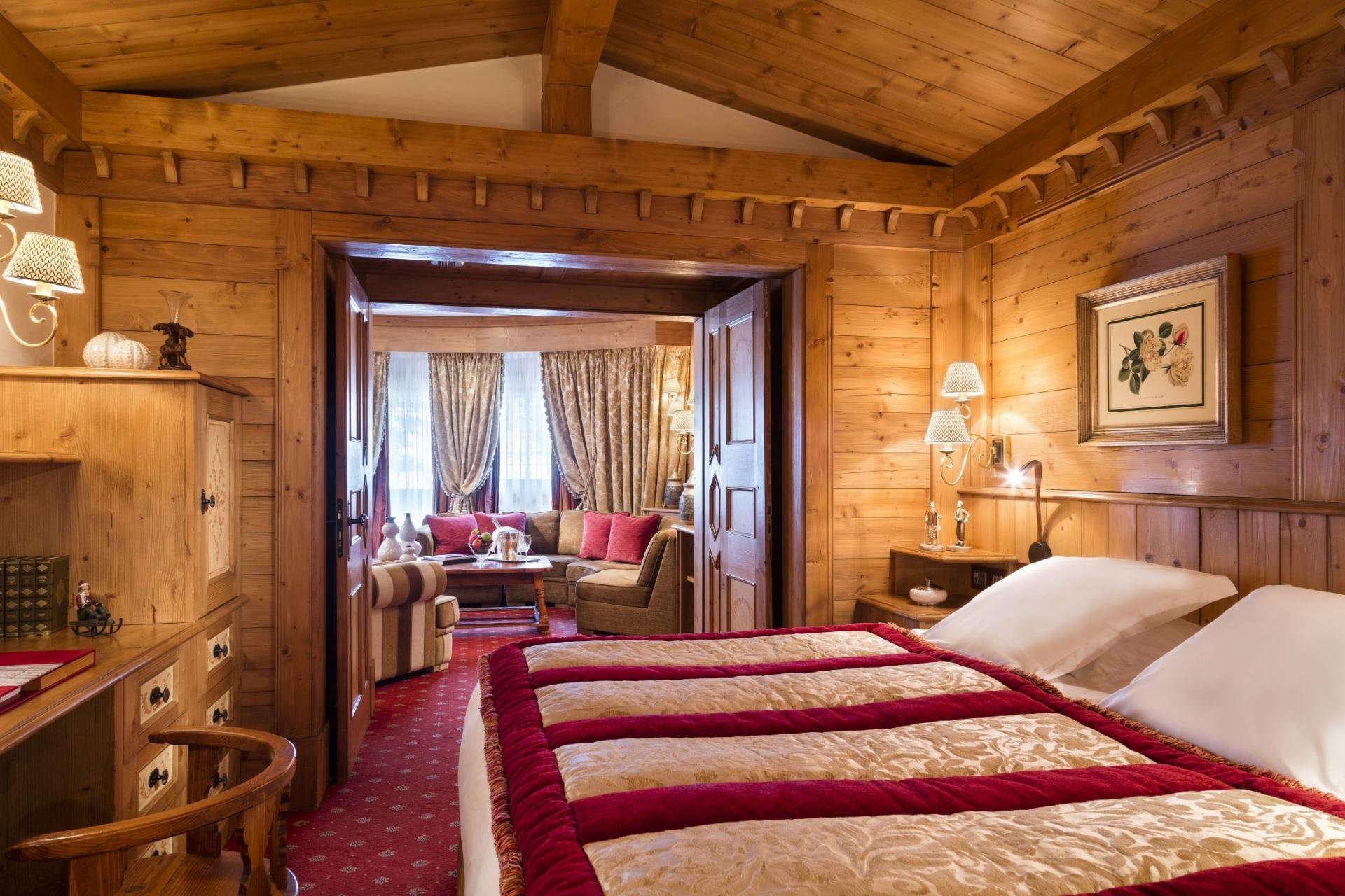 A bedroom with wooden walls and ceiling, two beds with beige and red bedding, side tables with lamps, and a framed floral picture on the wall. In the background, there's a sitting area with curtains and chairs, and a coffee table.