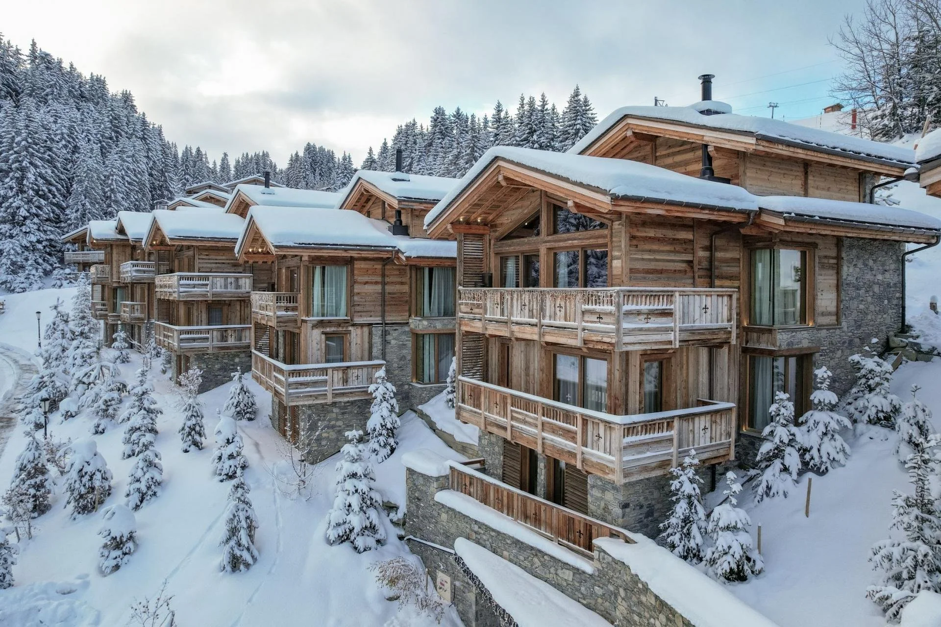 A row of wooden alpine-style chalets with snow-covered roofs, surrounded by snow-laden trees, in a mountainous winter landscape.
