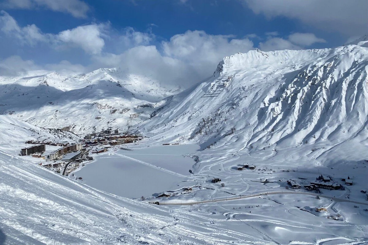 Snow-covered Tignes mountain landscape with a small town at the base, ski slopes, and a partially cloudy blue sky.