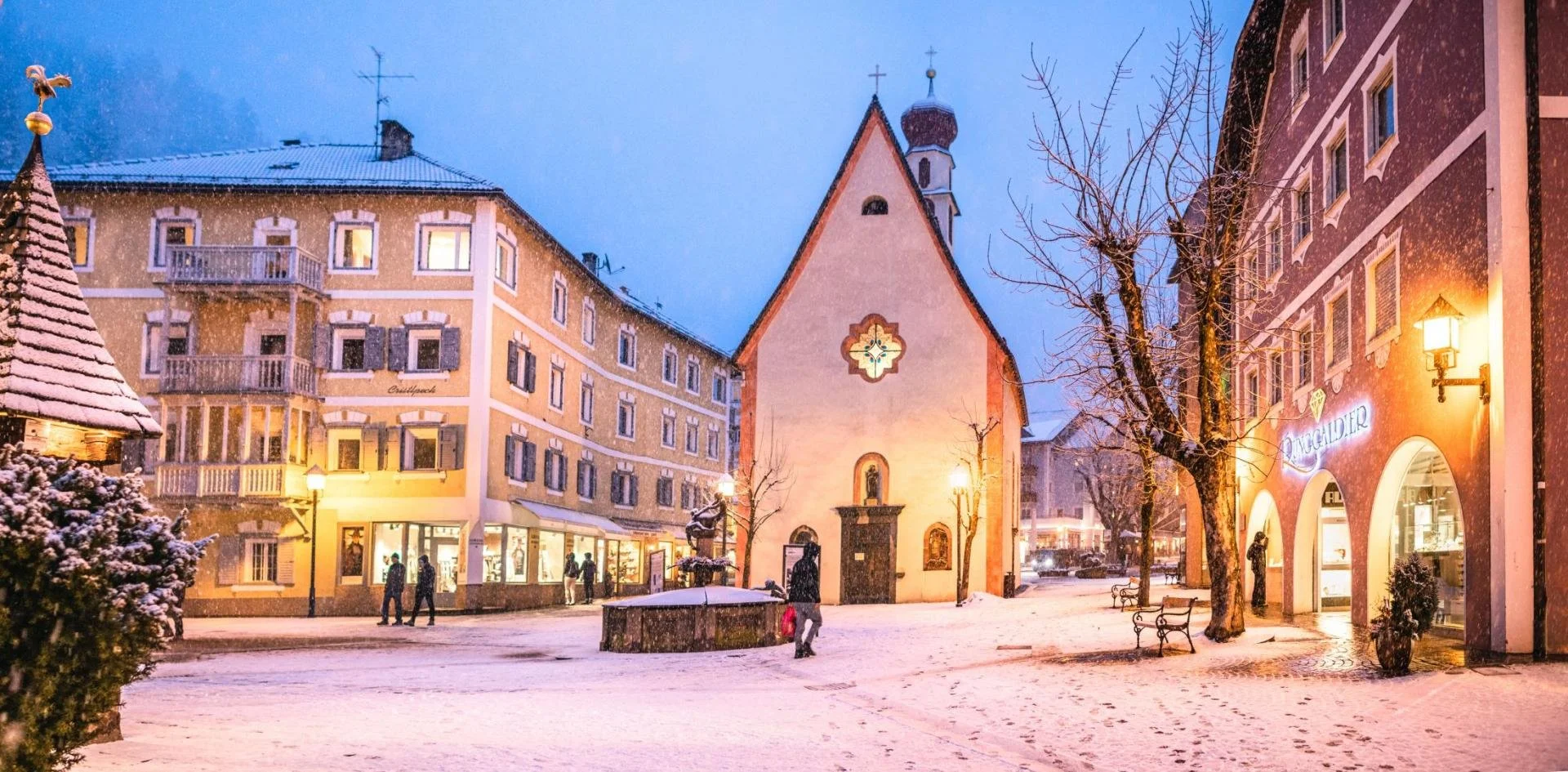 Snow-covered street with buildings, trees, and a small church in a European town at dusk, with streetlights on and people walking.