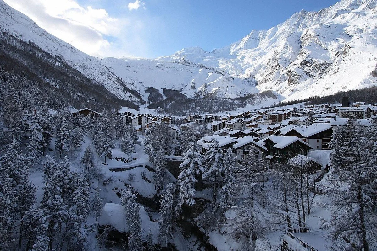 Snow-covered Saas-Fee alpine village with chalet-style buildings nestled among trees in a mountain landscape with snow-capped peaks in the background.