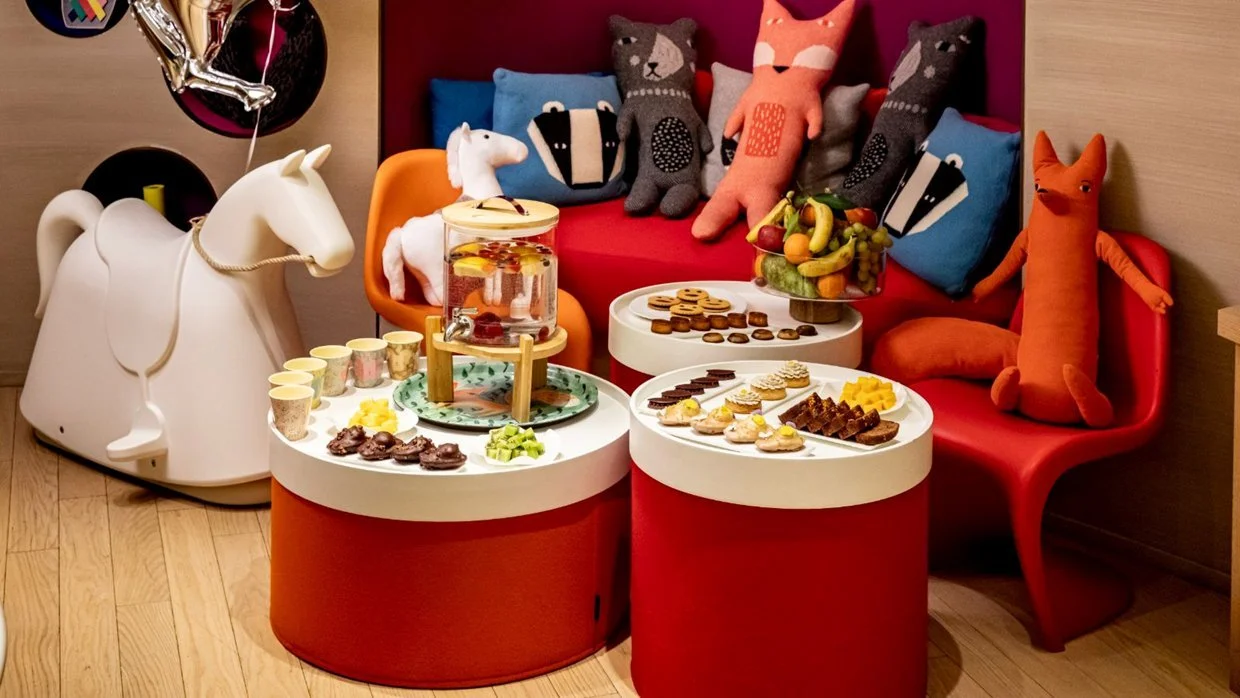 A children's birthday party setup with a white rocking horse, colorful plush toys, a small table with snacks and drinks, and a red couch with pillows and stuffed animals in the background.