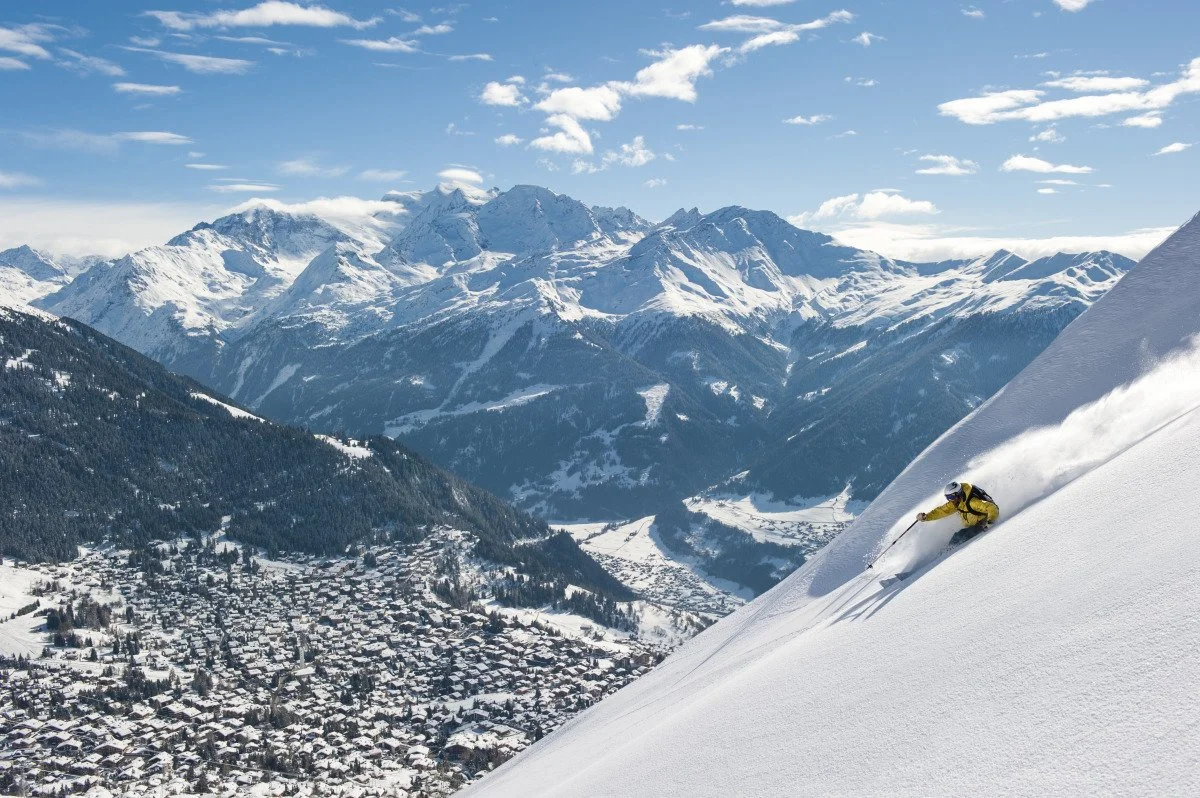 A skier in yellow gear skiing down a snowy mountain slope in Verbier with a village and snow-covered mountains in the background.