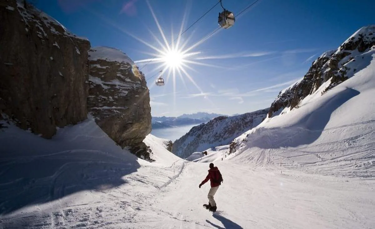 A person snowboarding down a snowy slope in Crans-Montana in a mountain valley with ski lifts, snow-covered peaks, and a bright sun in the sky.