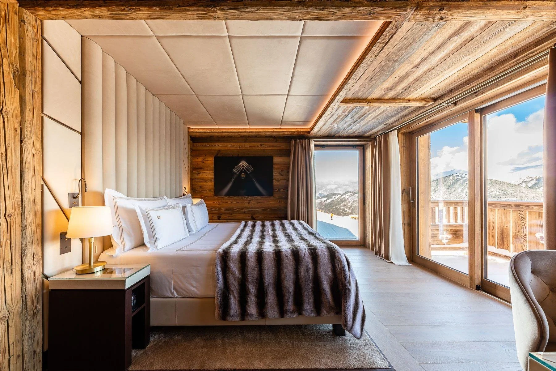 A cozy bedroom with a mountain view, featuring a large bed with white linens and pillows, a fur throw blanket, a bedside table with a lamp, and wooden walls and ceiling. There are large windows revealing snow-covered mountains outside.