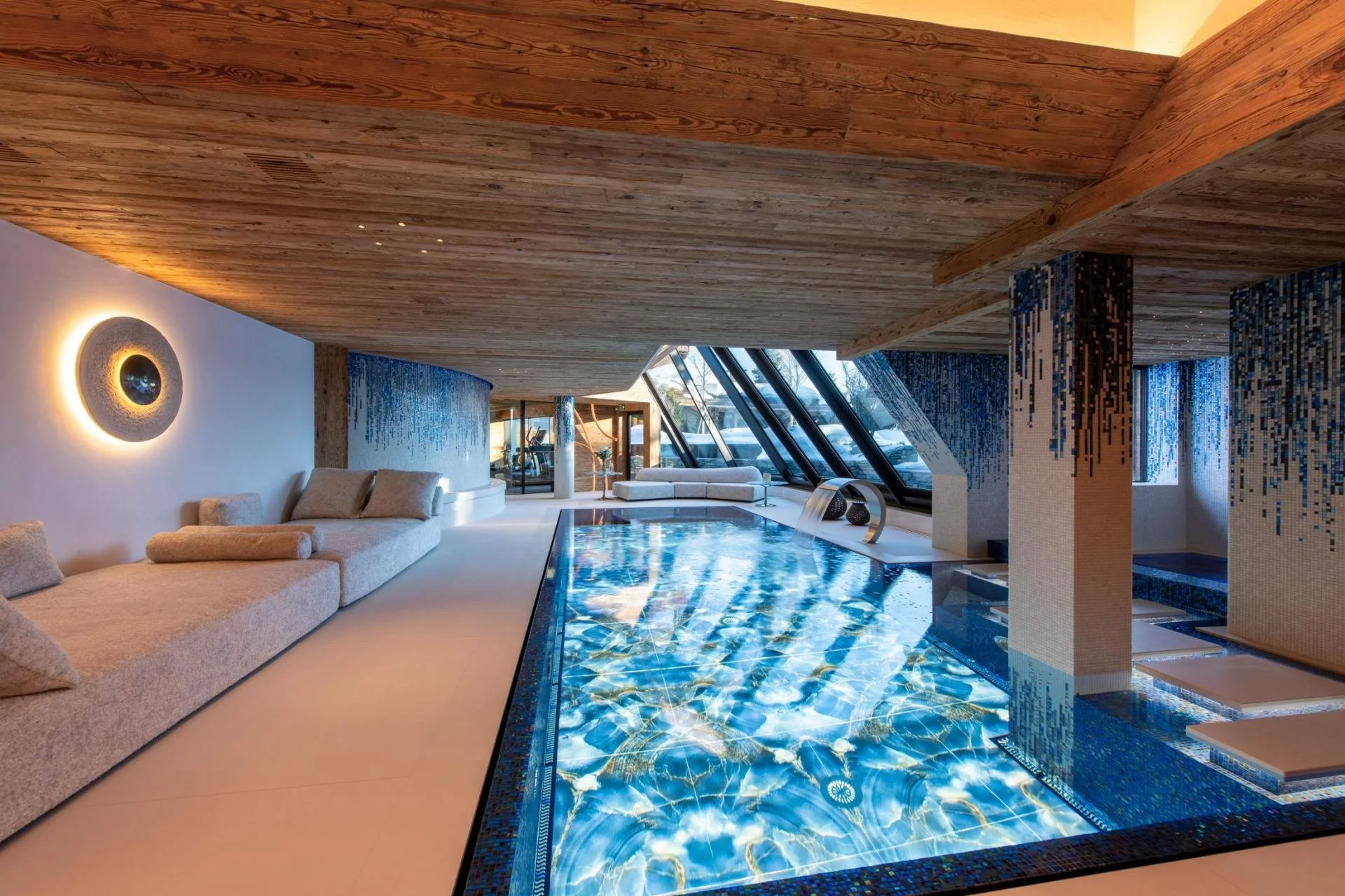 Interior of a modern house with a wooden ceiling, a beige sofa with pillows, a wall-mounted circular light, and a swimming pool with blue mosaic tiles under angled glass windows. There are decorative curtains and a small sitting area by the window.
