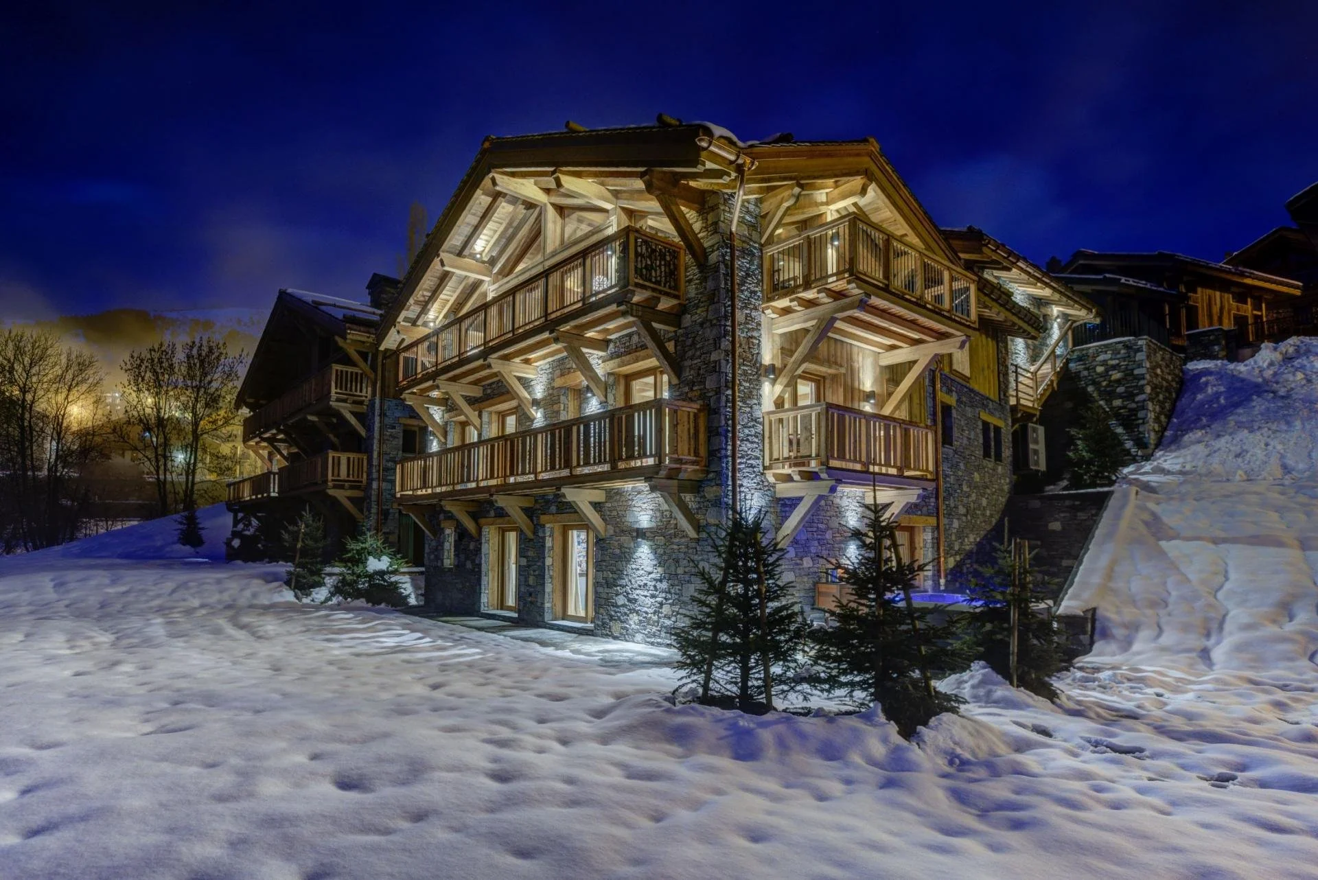 Nighttime view of a lit multi-story wooden and stone chalet-style house on a snowy slope with small evergreen trees and a dark blue sky backdrop.