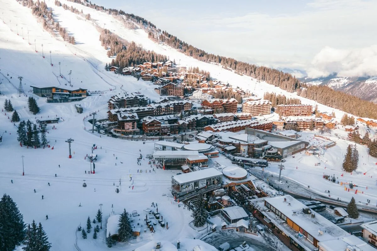 A snowy Courchevel 1850 with ski slopes, ski lifts, and a train station at the bottom, surrounded by trees and mountain peaks.