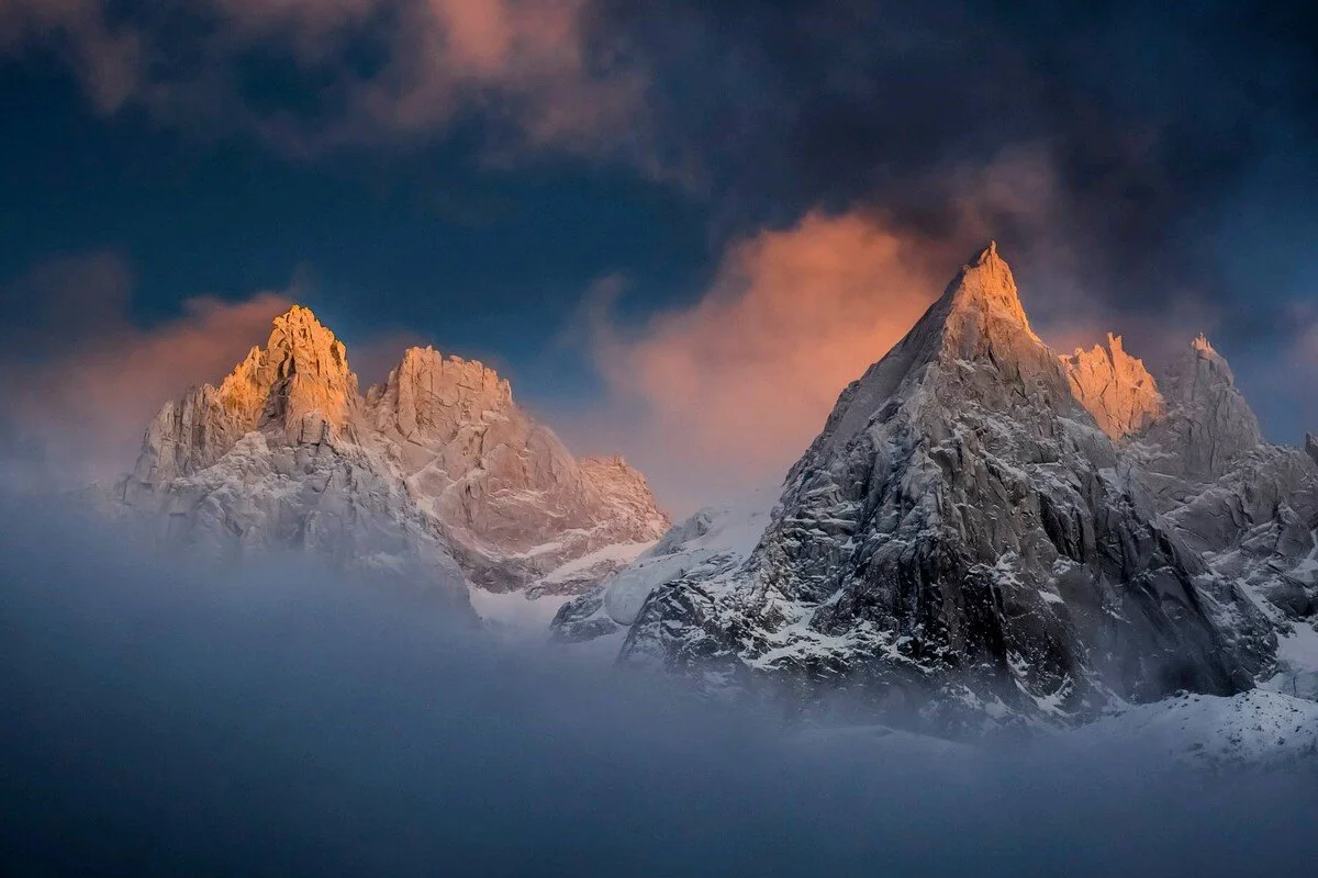Snow-covered mountain peaks in Chamonix illuminated by sunlight at sunset, with clouds surrounding the peaks.