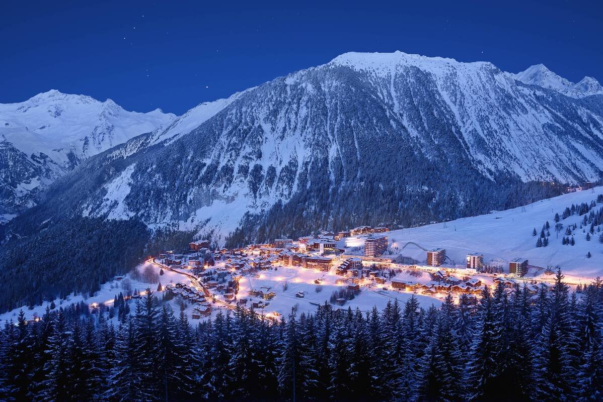 A snowy Courchevel 1650 village illuminated at night, with tall snow-covered trees in the foreground and a large mountain in the background.