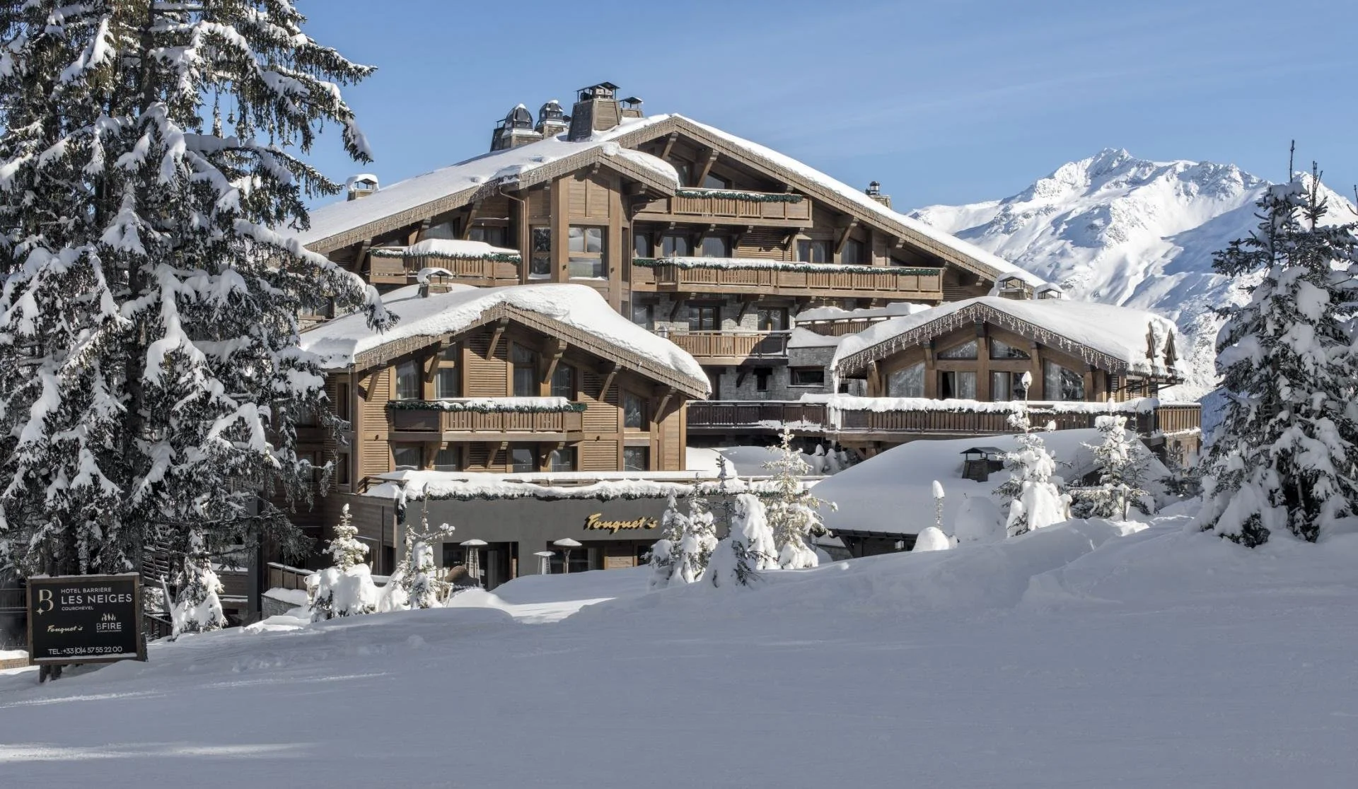 A large wooden chalet-style hotel covered in snow, surrounded by snow-covered pine trees and mountains, with a clear blue sky in the background.