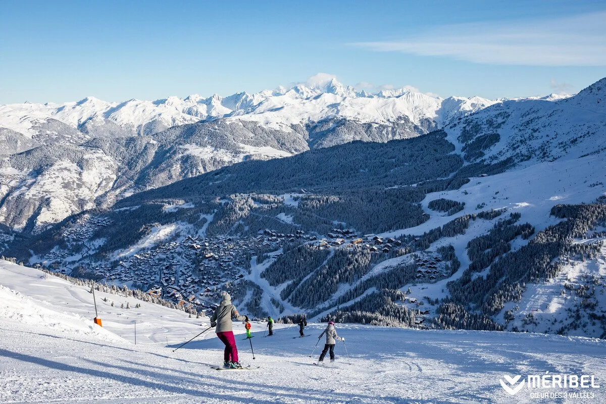 People skiing on a snowy mountain slope in Meribel with a village and snow-covered mountains in the background on a clear day.