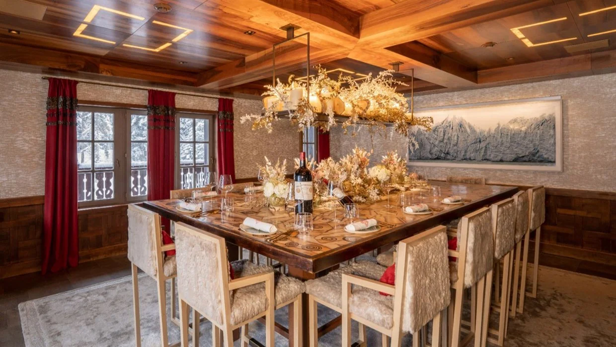 A beautifully decorated dining room with a wooden table set with wine glasses, plates, and napkins, and a large floral centerpiece. The room has red curtains, windows showing a snowy outdoor scene, and a textured wall with a landscape painting. Overh