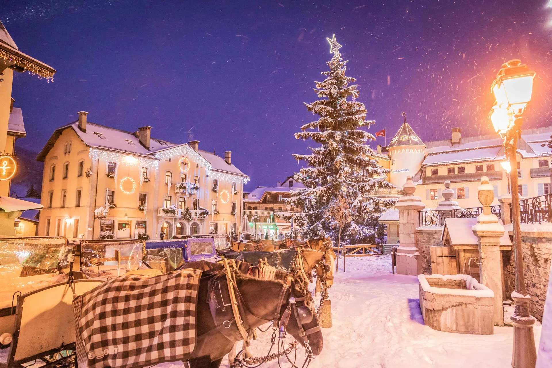 Snow-covered Megeve town square at night with decorated buildings, a large lit Christmas tree, horses hitched to sleds, and warm street lamps.