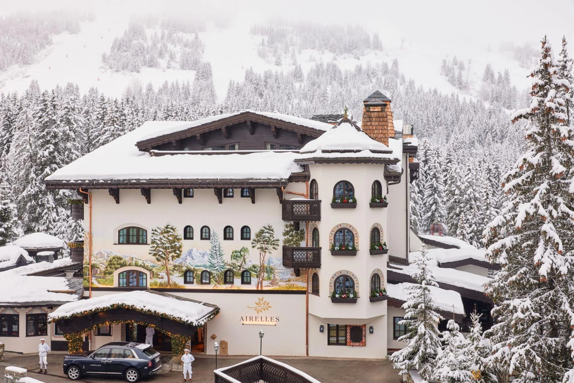 A large, elegant hotel with a white exterior and multiple balconies, surrounded by snow-covered trees and mountains, with decorative holiday lighting at the entrance and people in white uniforms standing outside.