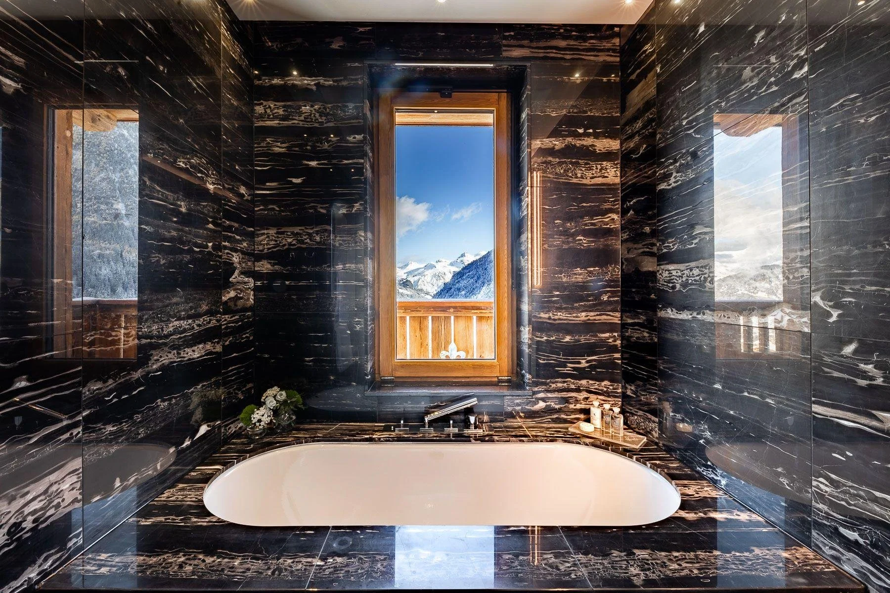 Luxurious bathroom with black marble walls and floor, a white oval bathtub, and a wooden window revealing snow-capped mountains and blue sky outside.