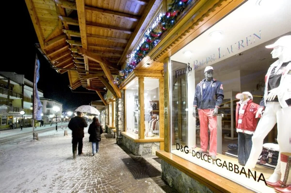 A street scene at night in Crans-Montana with two people walking under umbrellas in front of a store window display. The store features mannequins dressed in casual and winter clothing, with a sign indicating the brands D&G and Dolce & Gabbana. The s