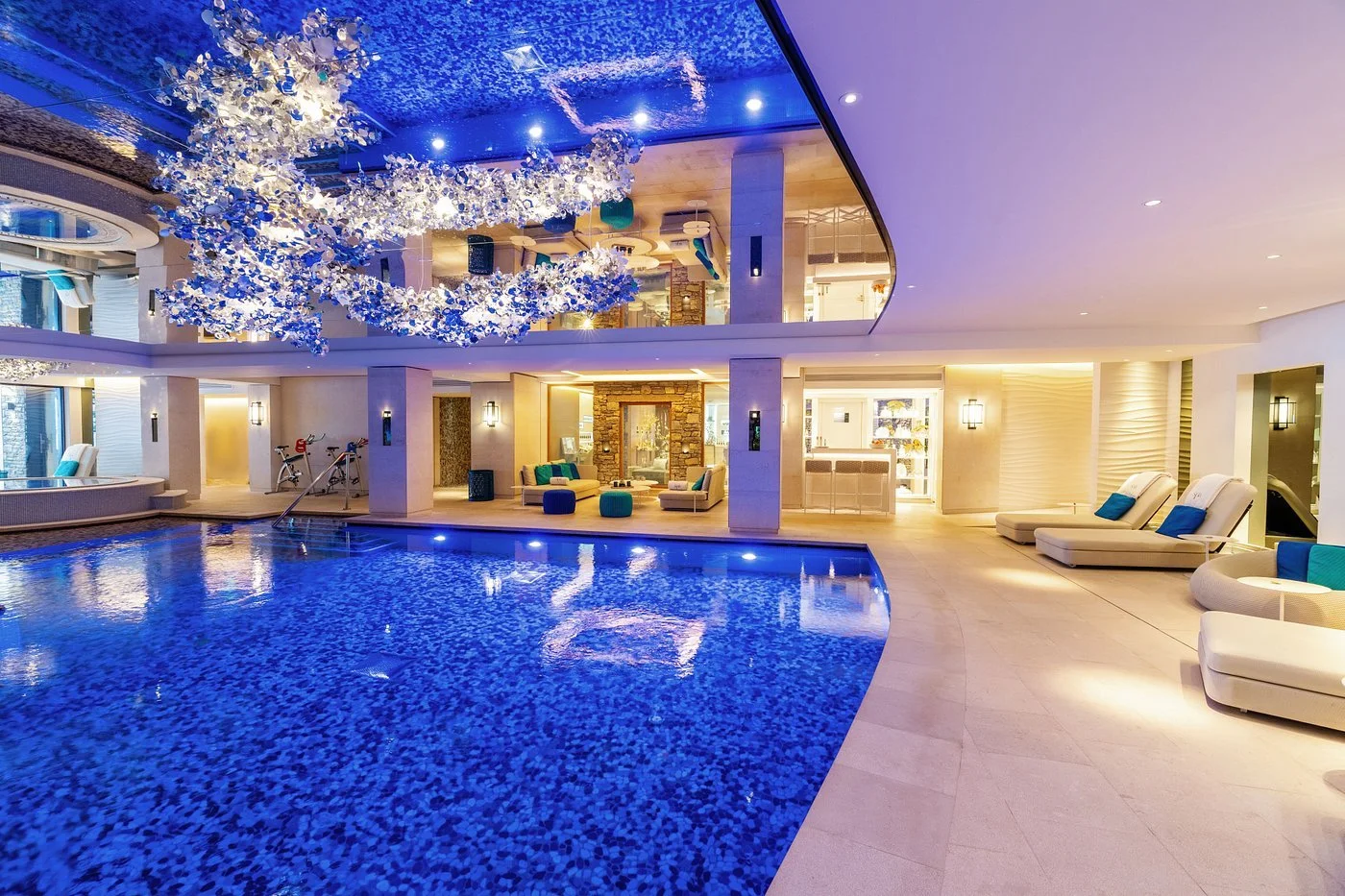 Luxurious indoor swimming pool area with lounge chairs, a decorative ceiling with hanging flowers, and modern lighting.