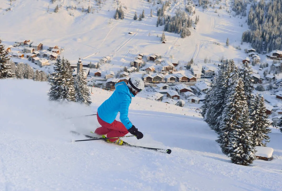 A skier wearing a blue jacket, red pants, black gloves, and a white helmet descending a snowy slope in Lech with a village and snow-covered trees in the background.