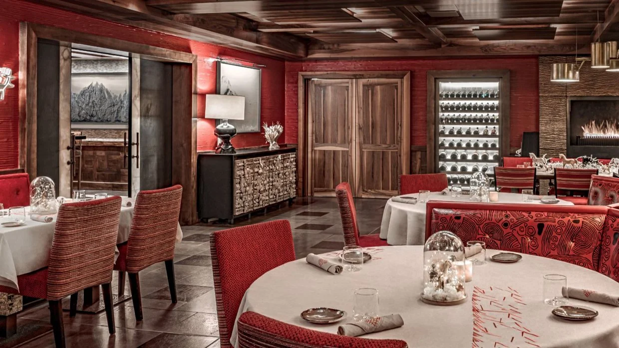 Elegant restaurant decorated in red and wood tones, with round tables set for dining, including glass cloches, candles, and rolled napkins, and a wine fridge featuring bottles of wine.