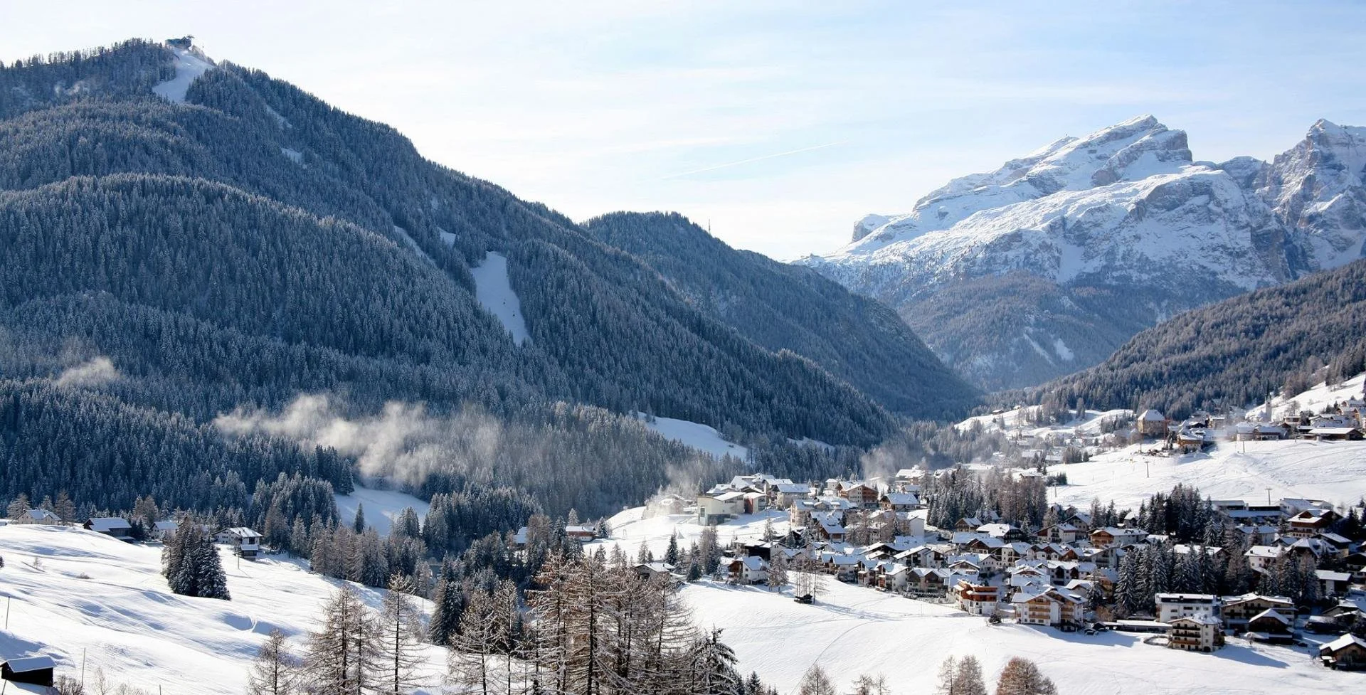 Snow-covered mountain village surrounded by dense forests and tall mountains under a clear blue sky.