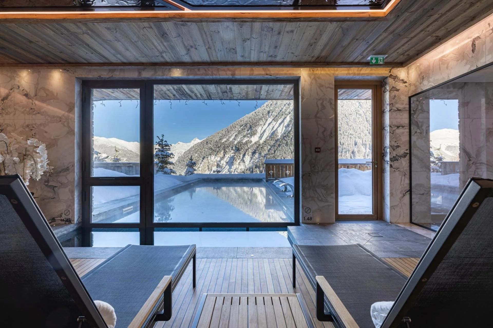 Indoor lounge area with wooden flooring and modern decor, overlooking a snowy mountain landscape through large glass windows and sliding doors.