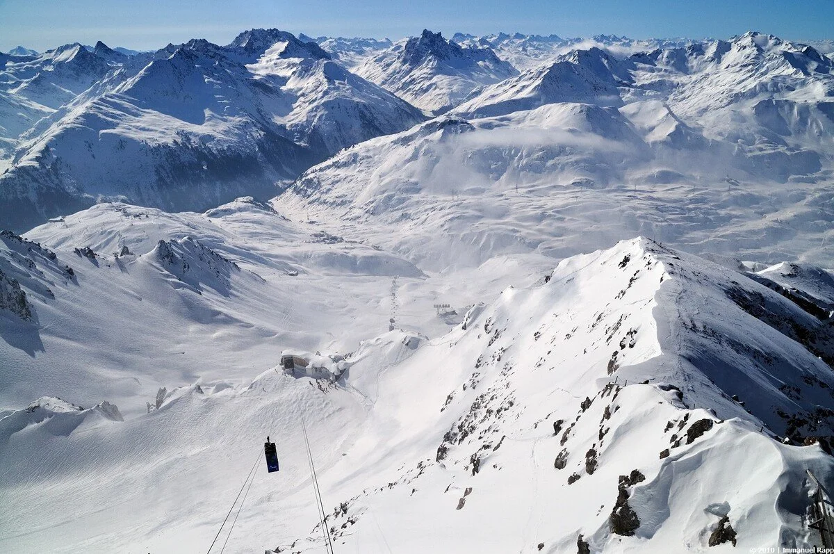 Snow-covered mountain peaks with a ski lift and ski tracks visible in the foreground.