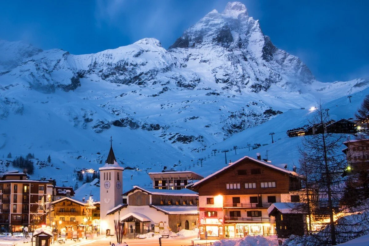 Snow-covered mountain village at dusk with chalet-style buildings, a church with a steeple, and ski lifts against towering snow-capped peaks.