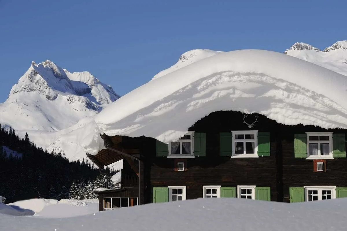 A wooden house in Lech with green shutters partially covered by a large snowdrift in a snowy mountain landscape.