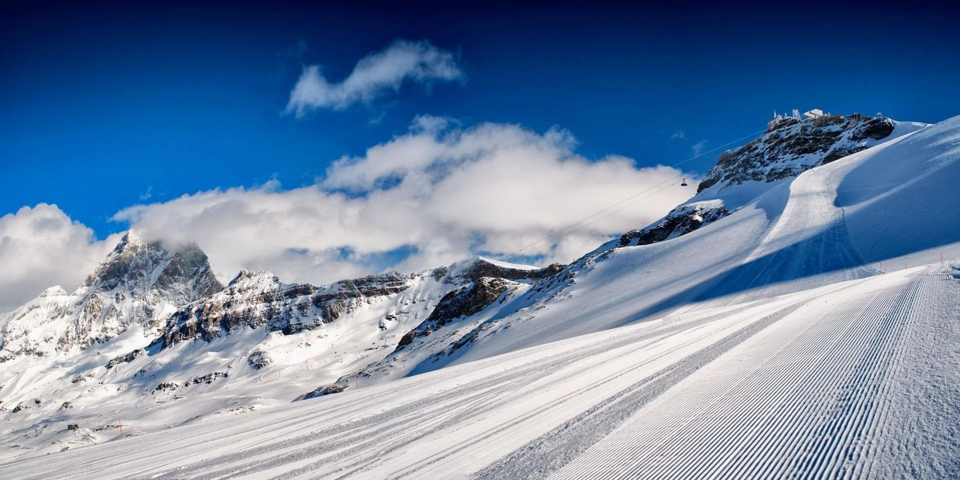 Snow-covered mountain slopes with groomed ski tracks under a partly cloudy blue sky. Ski lifts are visible on the right side near the mountain top.