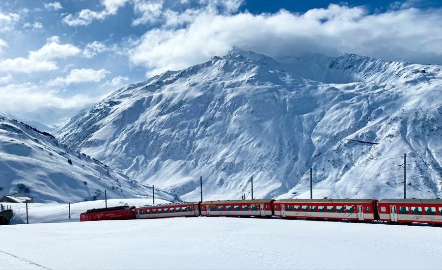 A red passenger train traveling through a snowy landscape in Andermatt with tall, snow-covered mountains in the background.