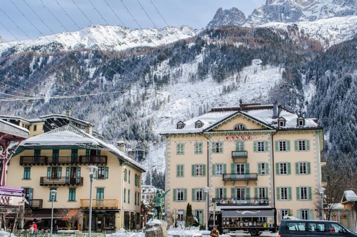 Snow-covered buildings in a Chamonix village with forested slopes and peaks in the background.