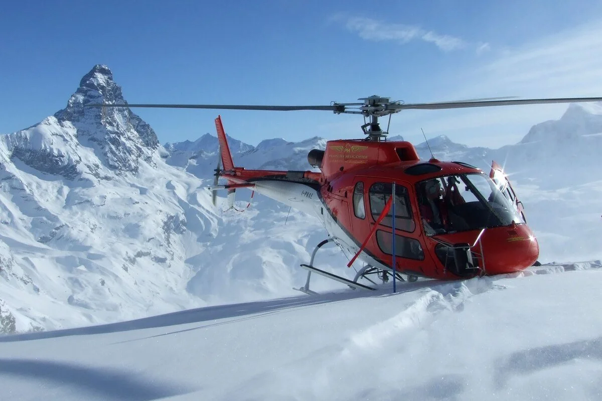 A red helicopter landed on snowy mountain terrain with a backdrop of snow-covered peaks and a partly cloudy blue sky.