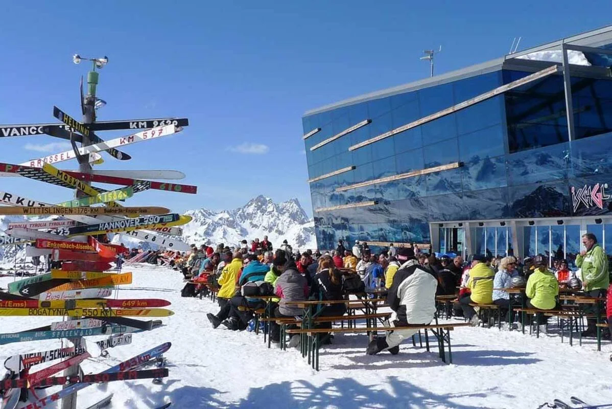 People sitting at outdoor tables in a snowy Ischgl mountain landscape, with a modern glass building on the right and snow-capped mountains in the background. A colorful signpost with numerous directional signs is on the left.