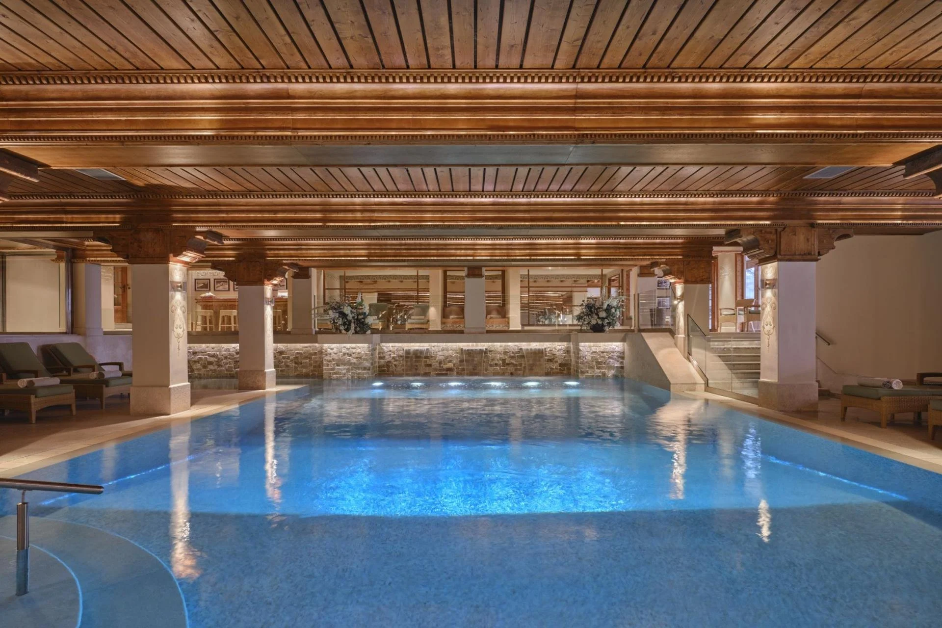 Indoor swimming pool with a wooden ceiling, lounge chairs, and a tiled wall with flowers on a balcony overlooking the pool.