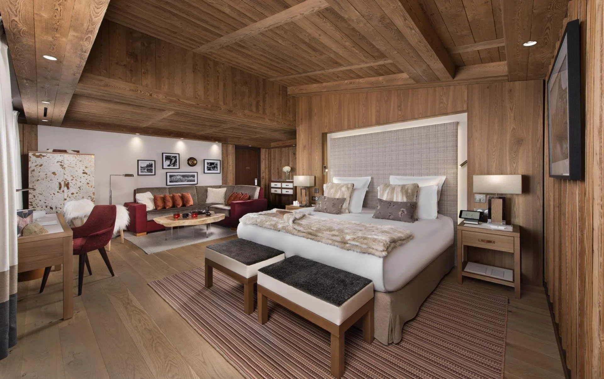 A cozy hotel room with wooden walls and ceiling, featuring a large bed with fluffy pillows and a fur throw, a sitting area with a sofa and armchair, and a wall decorated with black-and-white photos.