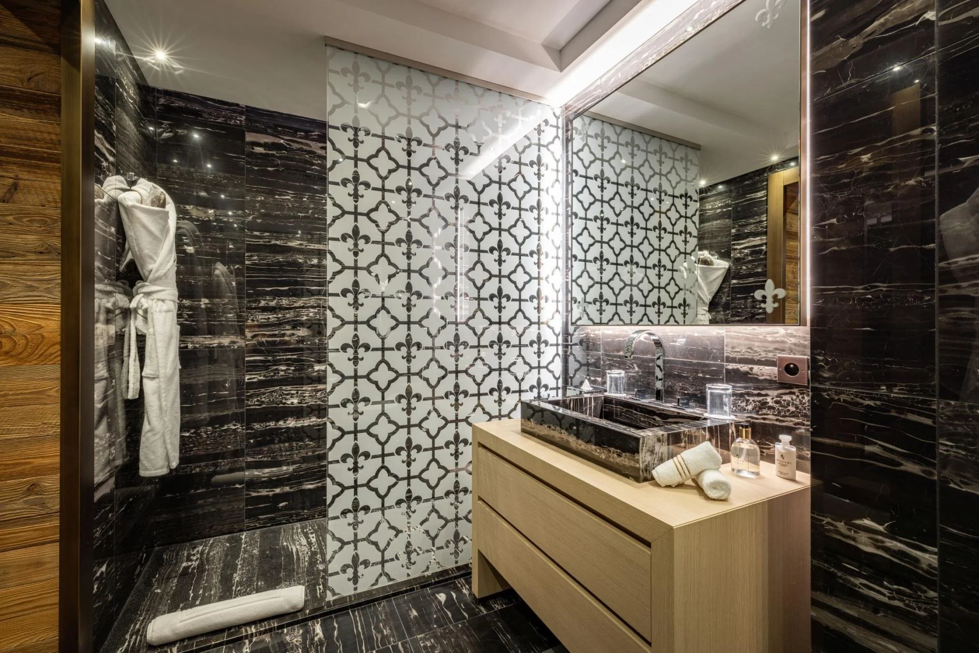 Modern bathroom with black and white patterned tiles, black marble walls, wooden accents, and a large mirror.