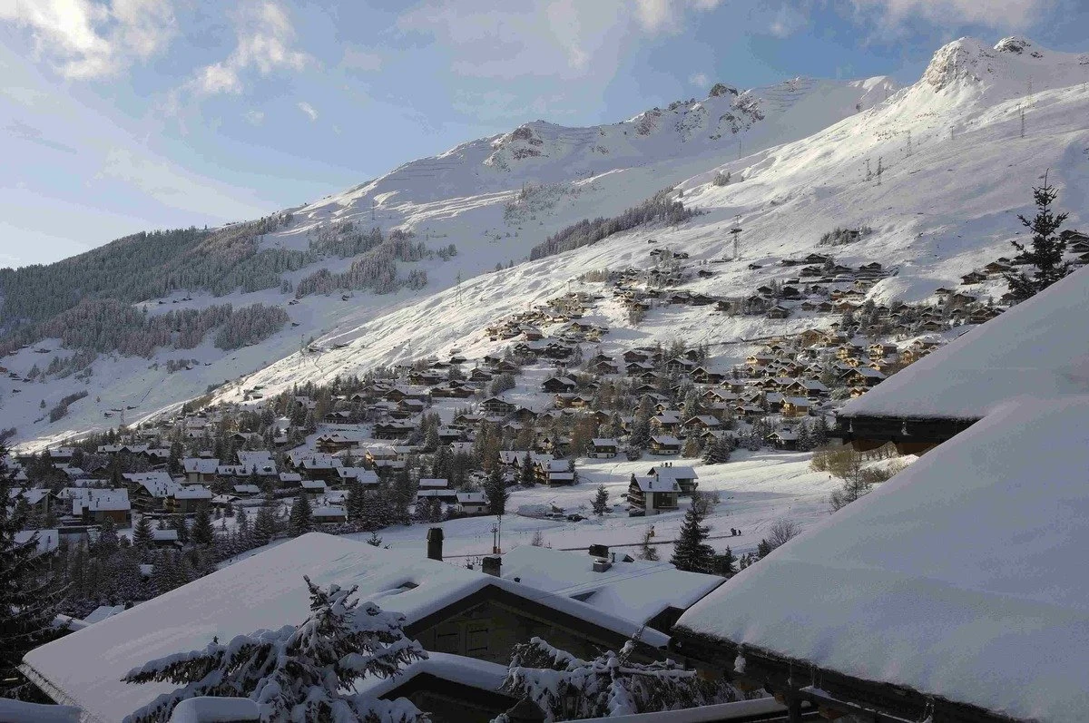 Snow-covered Verbier mountain village with scattered houses, ski lifts, and pine trees under a blue sky.