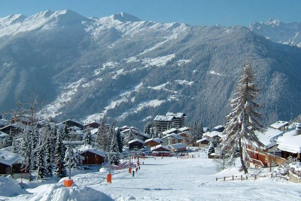 Snow-covered Verbier mountain village with ski slopes, surrounded by forested mountains under a blue sky.