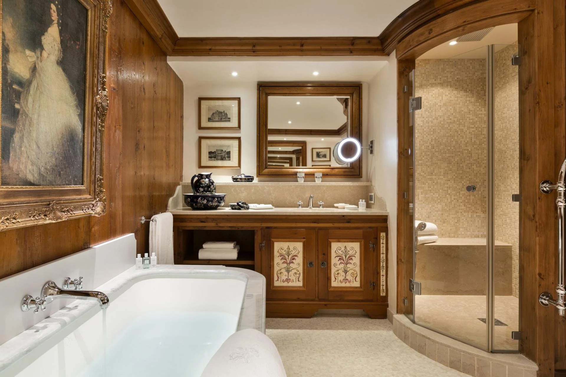 A luxurious bathroom with wooden accents, including a large mirror, decorative paintings, a bathtub filled with water, and a walk-in shower with a glass door.
