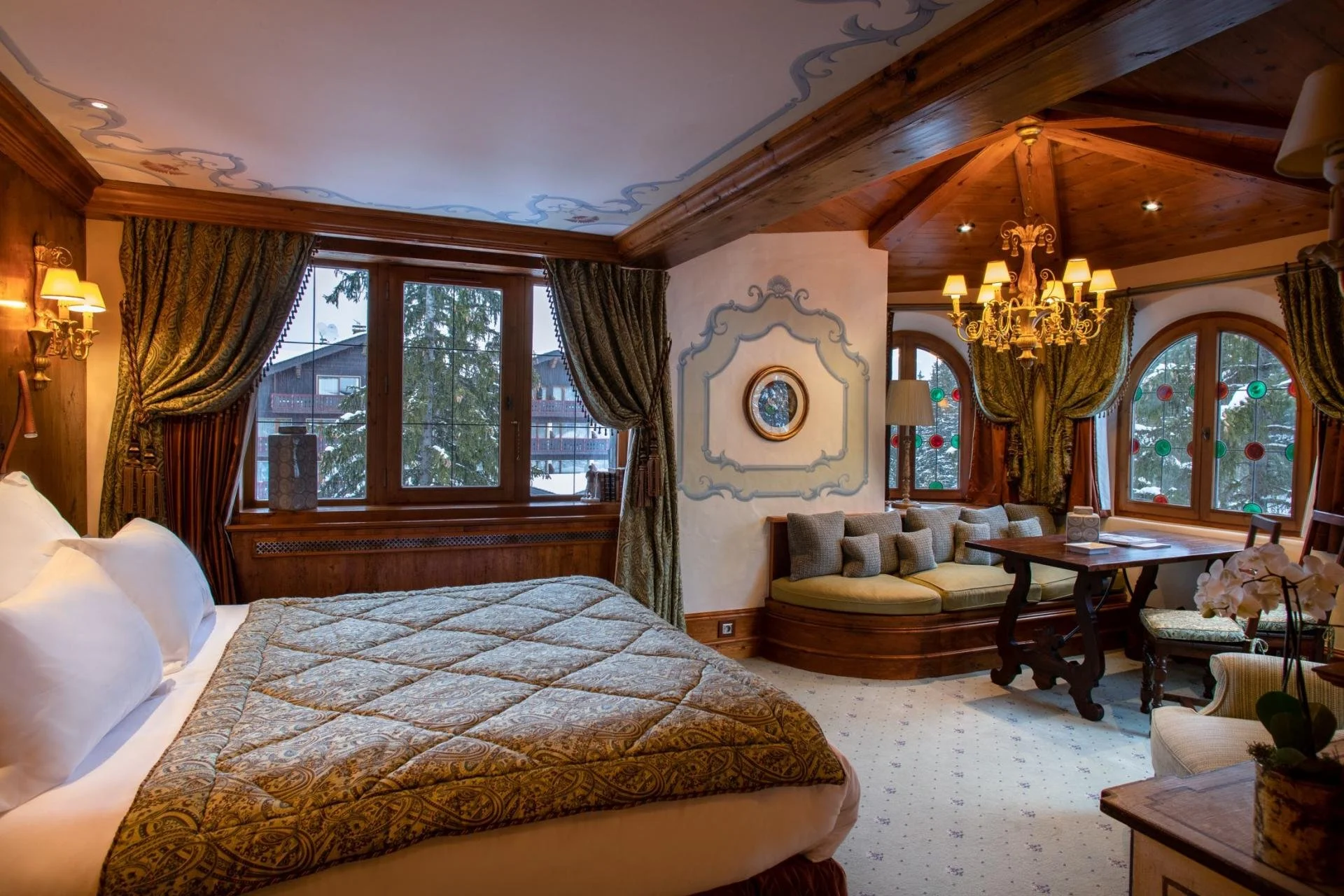 Luxurious bedroom with wooden ceiling beams, ornate chandelier, and bay windows with curtains, overlooking snowy trees and buildings outside.