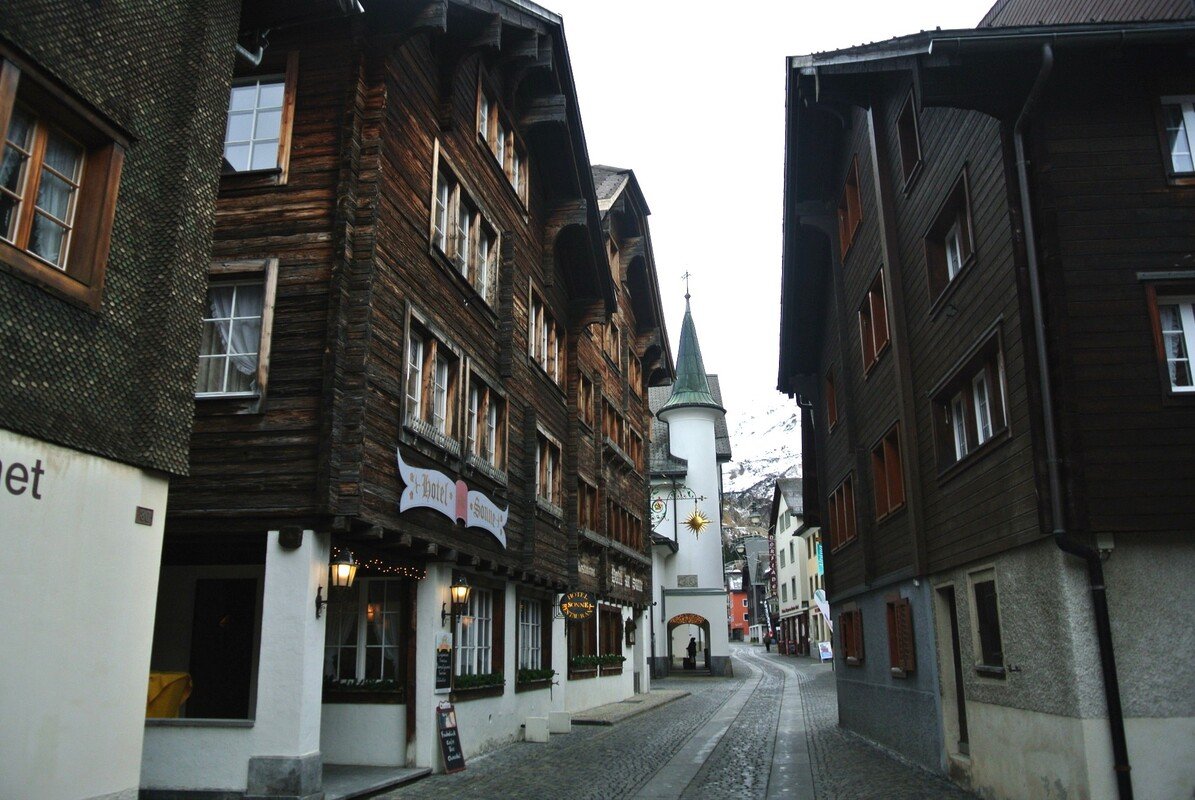 Narrow cobblestone street in Andermatt lined with dark brown and white buildings, featuring a church with a green spire in the background.