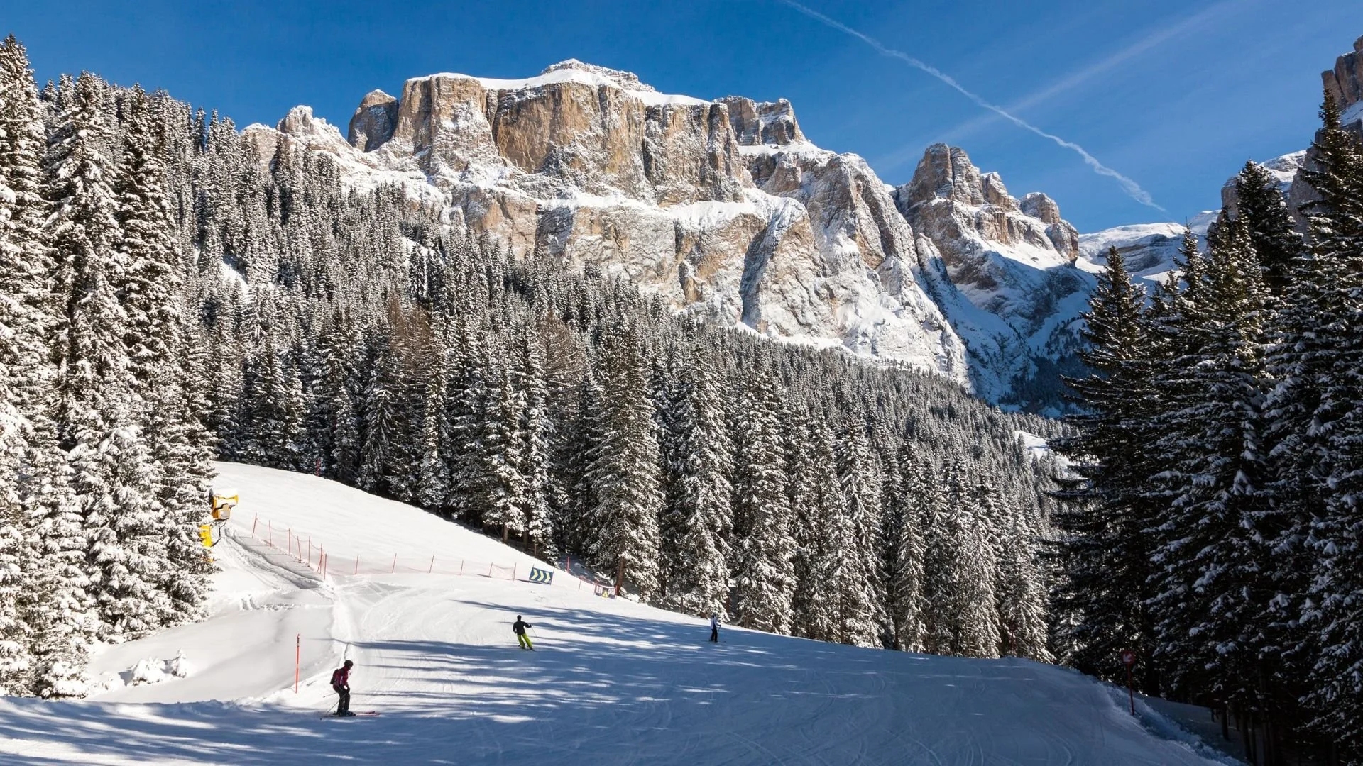 Snow-covered mountain landscape with dense pine forest and a ski slope with four skiers. Clear blue sky with some contrails.