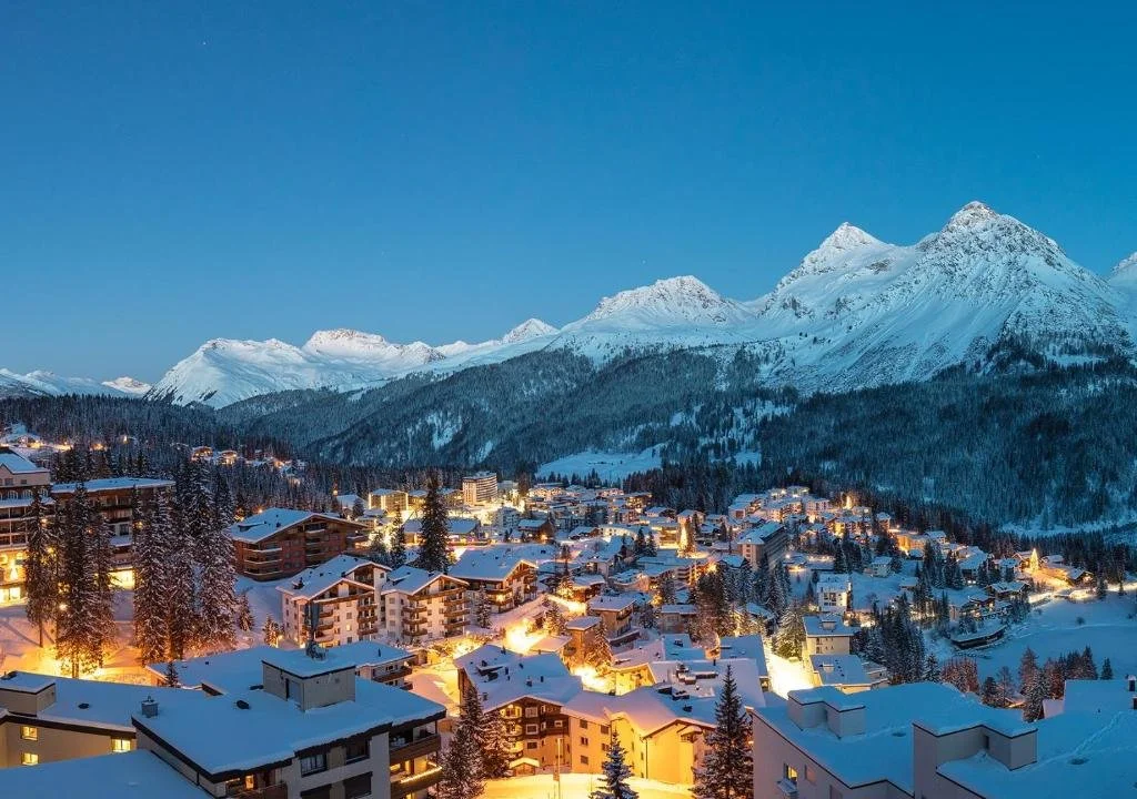 Snow-covered Arosa mountain village at twilight with lit windows and tall snow-covered trees.