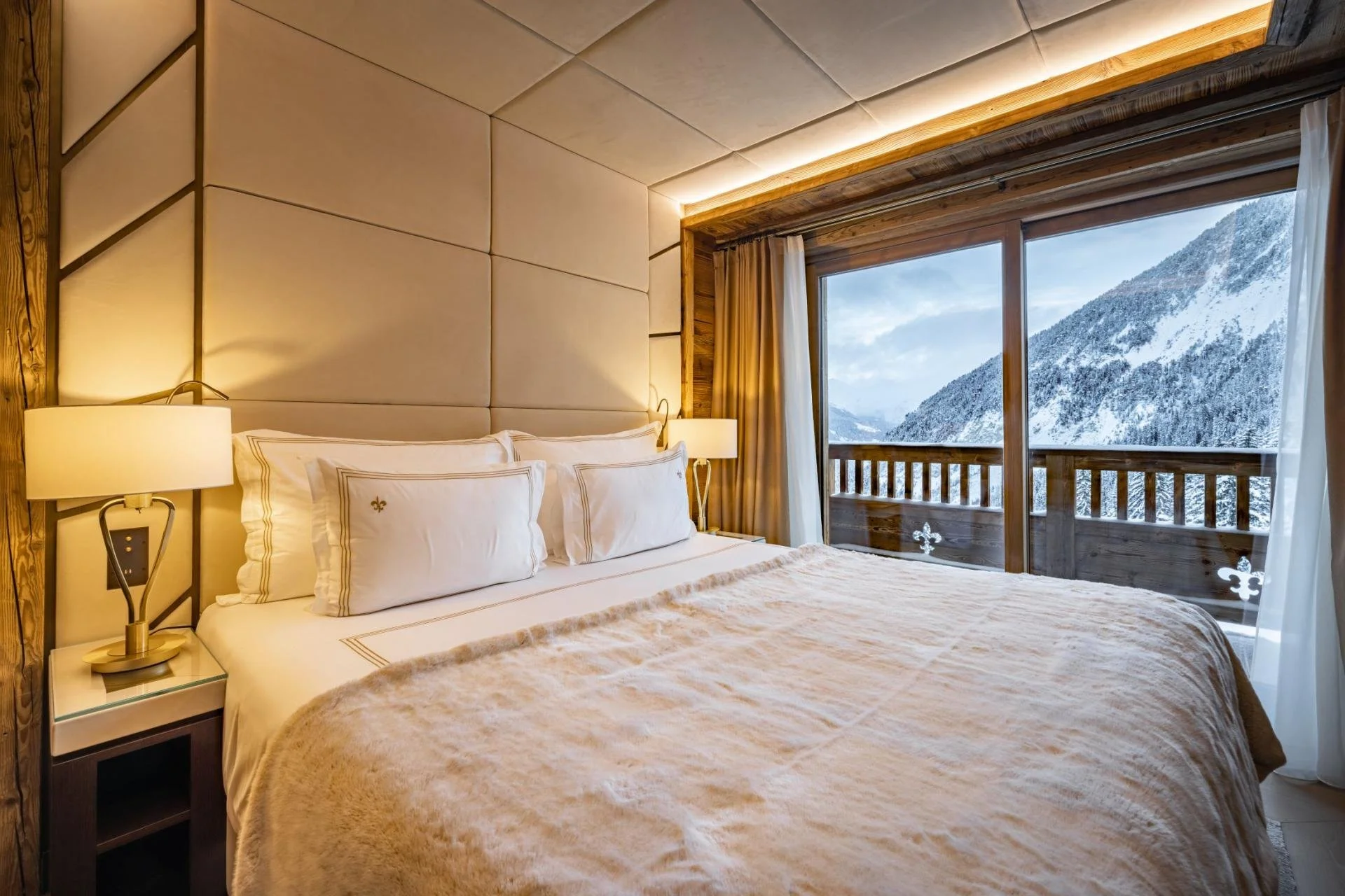 A cozy bedroom with a large bed, beige bedding, and white pillows featuring gold trim, glowing bedside lamps, and a wide window showcasing a snowy mountain landscape with trees.