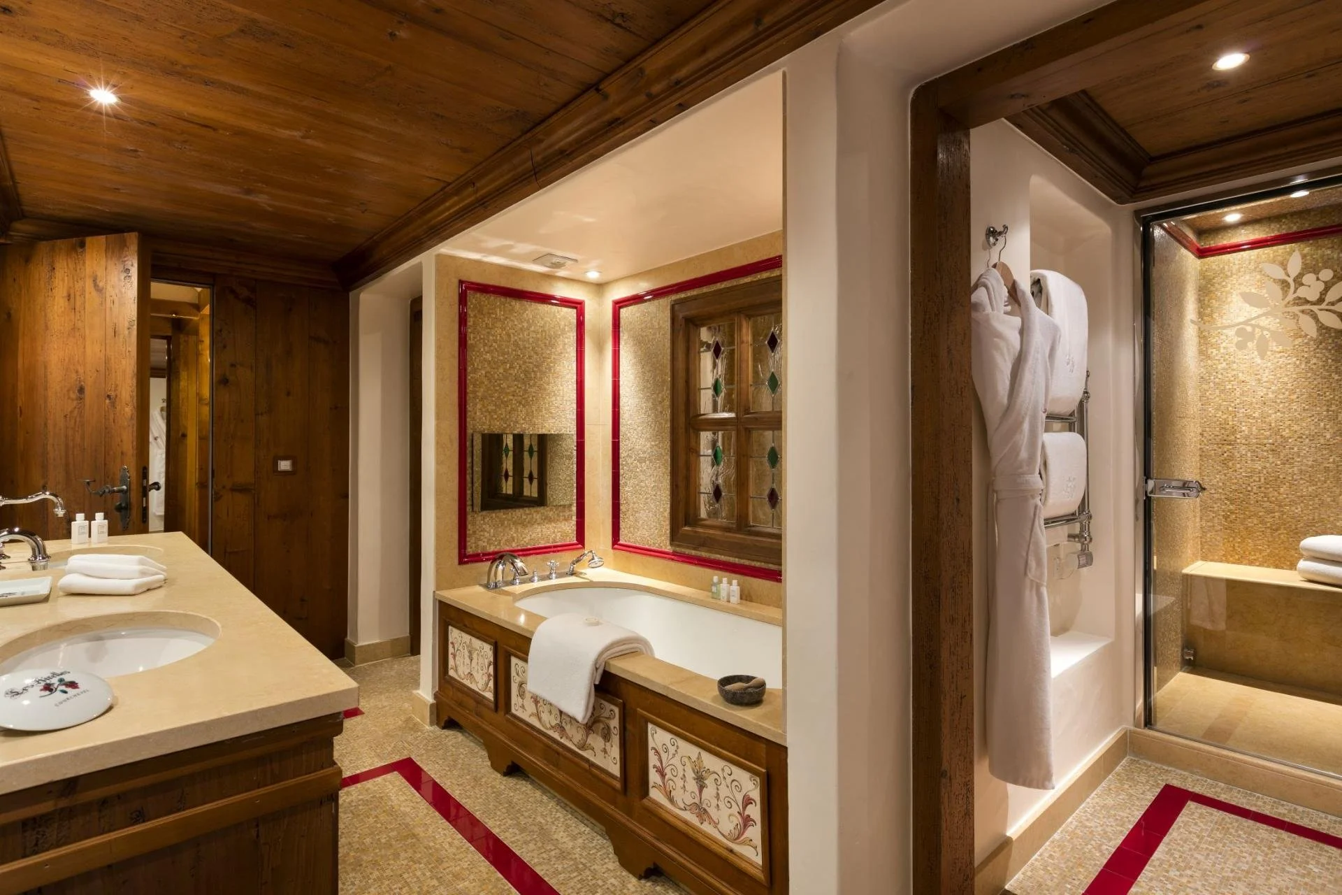 Interior of a luxurious bathroom with wooden ceiling and accents, featuring a bathtub, sink area, and a shower with glass door, decorated with gold mosaic tiles and red trim.